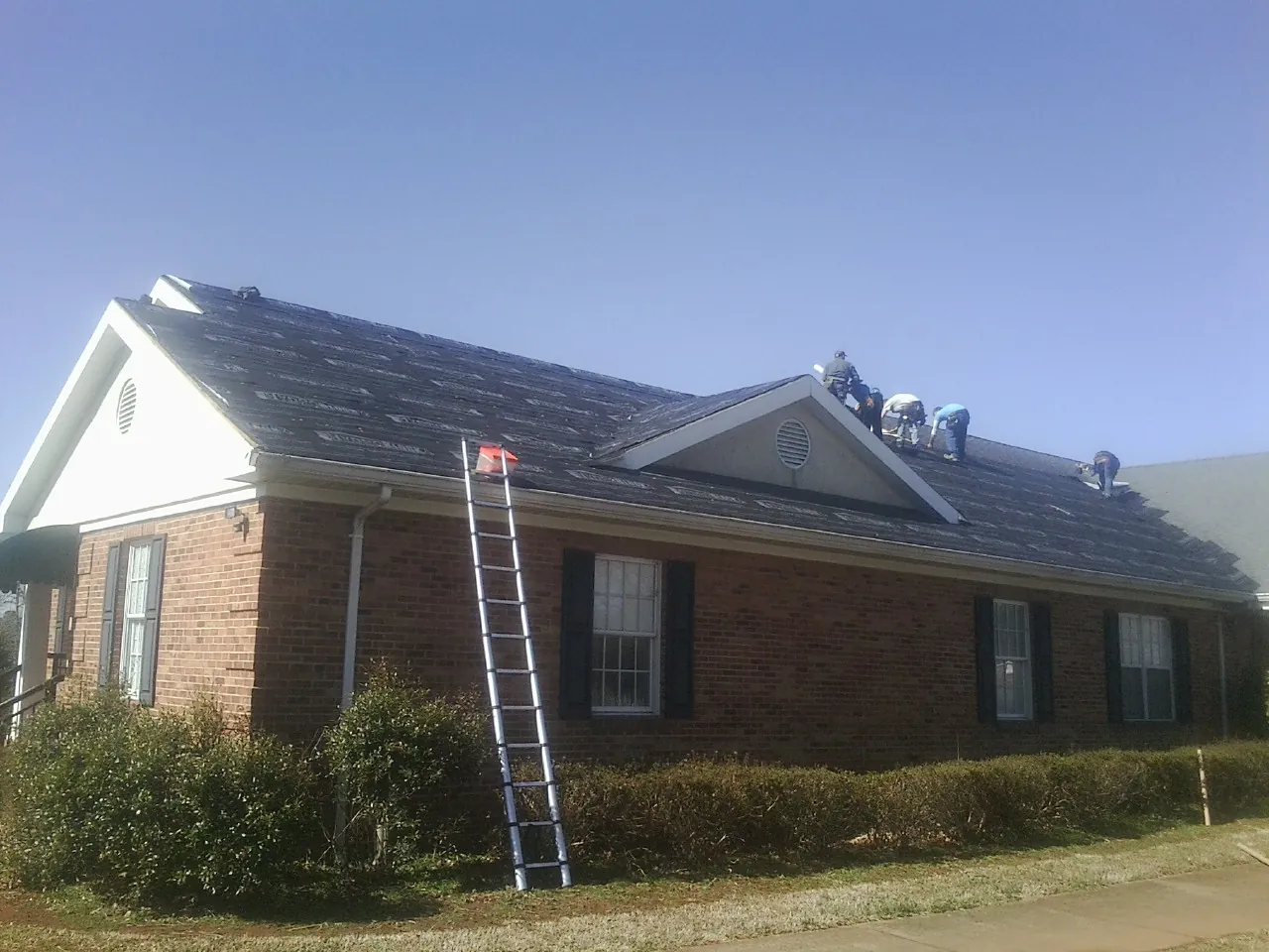 Crew tearing shingles off a brick ranch roof with a ladder set — Metal Roof Repair work by our crew in Letohatchee